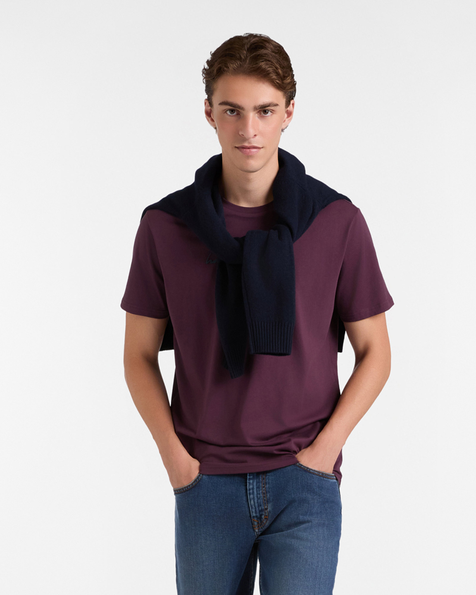 Man in plain purple t-shirt, blue jeans and dark blue sweater over shoulders